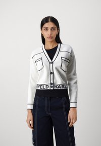 Vit croppad kavaj med svarta detaljer, med V-ringning, två fickor och en logoband i nederkanten. Mjuk textur och knäppning med knappar.