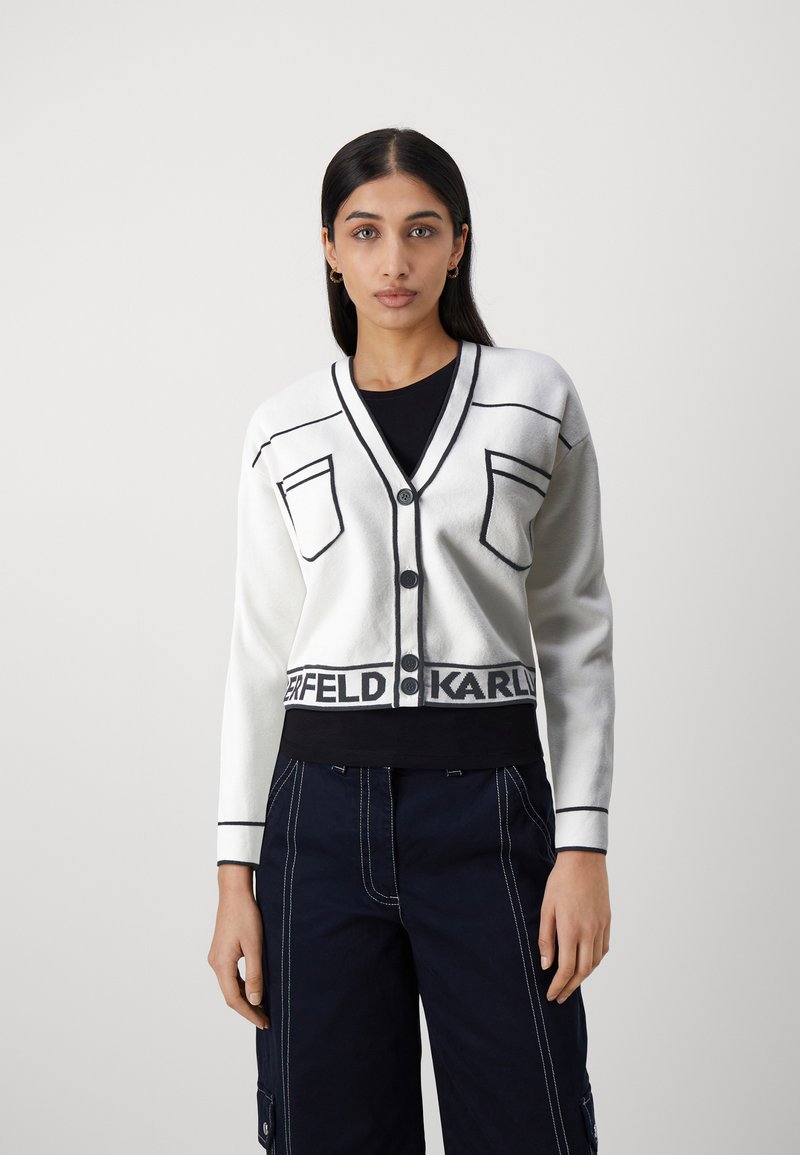 Vit croppad kavaj med svarta detaljer, med V-ringning, två fickor och en logoband i nederkanten. Mjuk textur och knäppning med knappar.