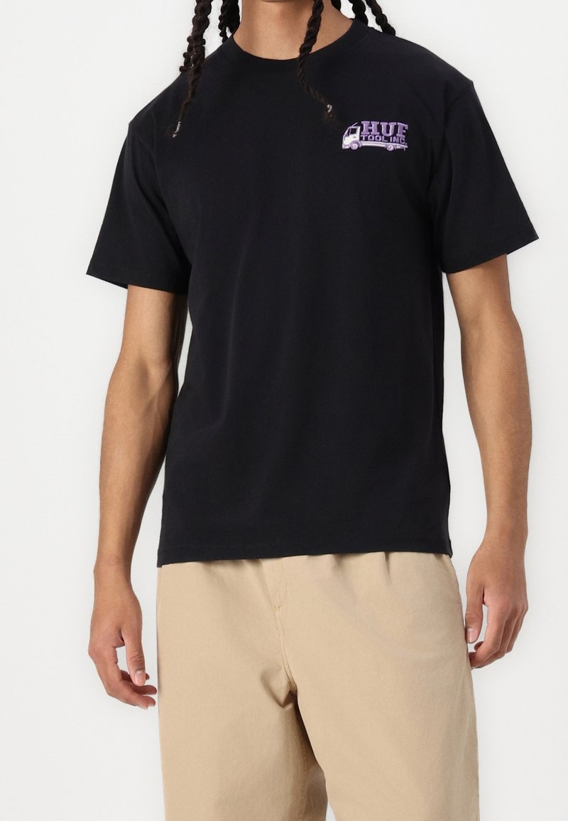 Homme portant un T-shirt noir avec un petit logo de camion violet et un pantalon beige, debout devant un fond blanc.
