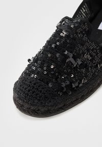 Svart espadrille med ett ovansidan täckt av paljetter, flätad jute-sula och en texturerad svart tygkant runt öppningen.
