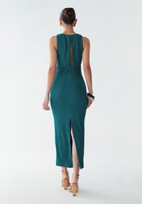 Žalsvai mėlyna megzta maksi suknelė su atvira nugara, šoniniu plyšiu ir aptempiančiu siluetu. Papildyta neutraliomis blokinėmis aukštakulnėmis bei apyranke.