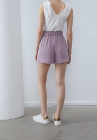 Haut blanc sans manches avec détails à volants ; short lilas avec taille élastique ; modèle portant des baskets blanches avec des accents dorés.