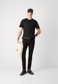Maglietta nera a girocollo con logo discreto, jeans neri aderenti, scarpe nere e un cappellino nero; tiene in mano una giacca chiara.