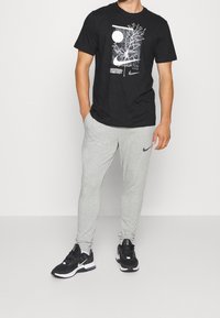 Man i svart grafisk Nike-t-shirt, ljusgrå Nike sweatpants och svarta Nike sneakers står med händerna i fickorna.