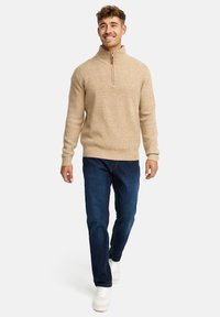 Homme marchant, portant un pull zippé beige, un jean bleu foncé et des baskets blanches, sur un fond clair uni.