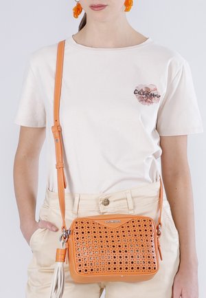 Vrouw draagt beige broek en wit T-shirt met bloemlogo, met een oranje crossbodytas met geperforeerd ontwerp en witte kwastdetail.