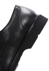 Chaussure en cuir noir avec une texture lisse, des coutures contrastantes, un talon légèrement surélevé et un design à bout arrondi.