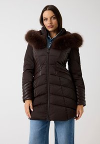 Cappotto imbottito marrone con cappuccio in pelliccia sintetica removibile, texture trapuntata, zip frontale e tasche laterali, abbinato a jeans blu.