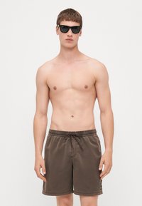 Brune svømmedrakter med elastisk liv og snor. Stoffet er glatt med en subtil glans og en liten logo på venstre side.