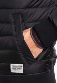 Musta, puffattu takki, jossa edustatasku ja merkki. Hihansuu on tumman viininpunainen neulekuvio, jossa on harmaa reuna.