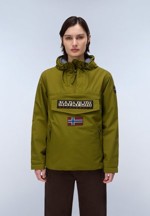 Personne aux cheveux courts et foncés portant une veste à capuche vert olive avec le logo Napapijri et un patch du drapeau norvégien, debout sur un fond uni.