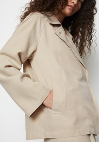 Beež jakk kergest kangast, laia krae, langenud õlgade ja küljetaskutega. Sile tekstuur koos õmmeldud detailidega.