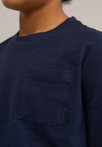 Jūras zila t-krekls ar apaļu kakla izgriezumu, kuram ir teksturēta kabata kreisajā krūtī un melna izšūta "R" logo.