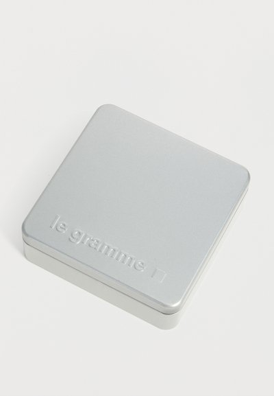 Boîte métallique carrée argentée avec coins arrondis étiquetée "le gramme" sur un fond blanc.