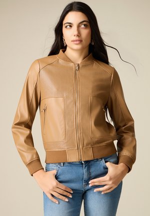 Femme aux longs cheveux foncés portant une veste en cuir perforé couleur camel avec fermeture éclair frontale et un jean bleu clair, debout les mains sur les hanches.