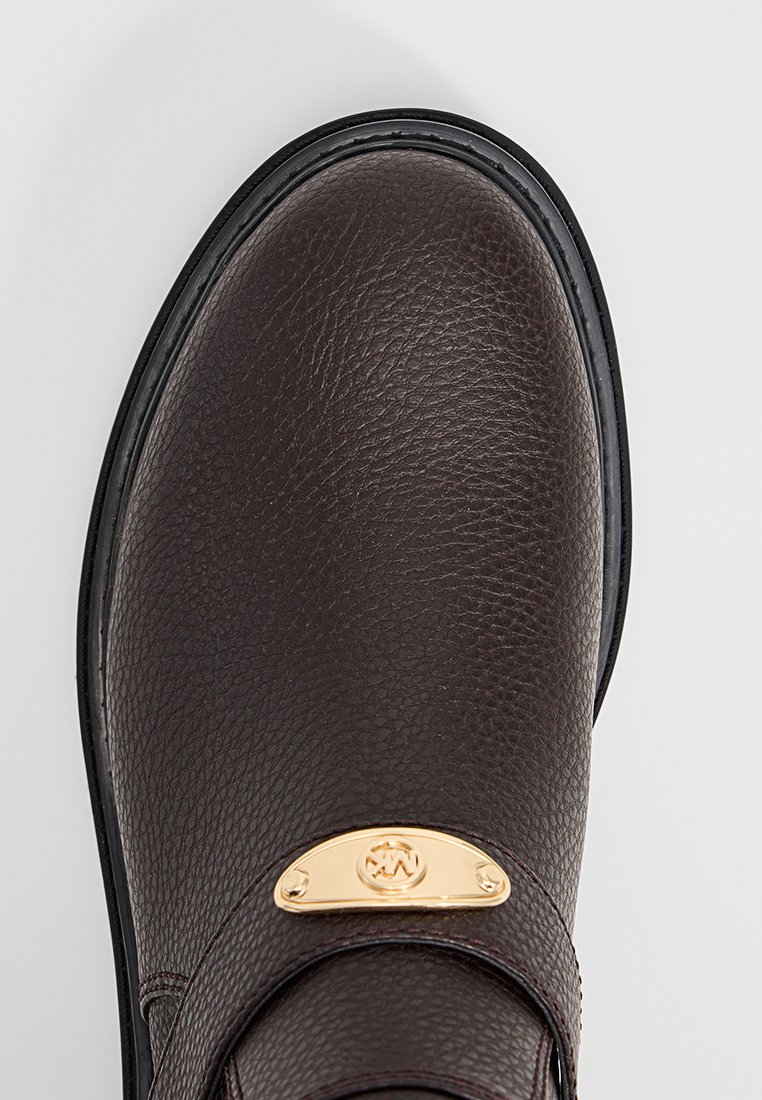 Zapato de cuero marrón oscuro con una superficie texturizada, que presenta una correa ancha y un acento de hardware en tono dorado. Suela de goma con borde negro.