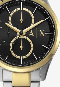 Ura s črnim številcem z zlatimi poudarki, ki ima srebrn pašček iz nerjavečega jekla, kronografske pod številčnice in izrazite oznake ur.