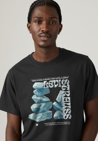 Μαύρο βαμβακερό μπλουζάκι με γραφικό σχέδιο από στοιβαγμένες γαλάζιες πέτρες και λευκό κείμενο που αποδίδεται στον Levi Strauss, στο κέντρο του μπροστινού μέρους.