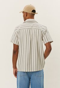 Kortslepet skjorte i beige med vertikale grønne striper. Har en nedbrettbar krage og ryggstykke. Laget av et lett materiale.