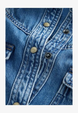 Farmering ing kék mosással, fém patentokkal és kontrasztos varrással. A közeli felvétel felfedi a textúrát és a különböző anyagot.