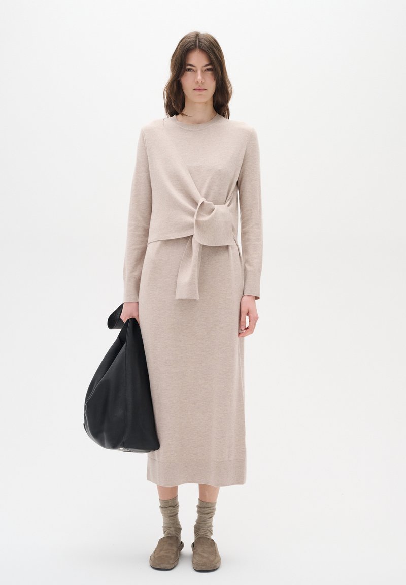 Béžové pletené šaty s designem do zavinování a uzlovým detailem, kombinované s černou koženou taškou. Modelka nosí taupe espadrilky a světlé ponožky.