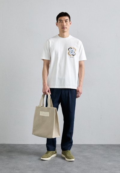 Hvid bomulds T-shirt med blomsterbroderi, navy bukser, grøn ruskinds sneakers og en beige tekstureret strandtaske med håndtag.