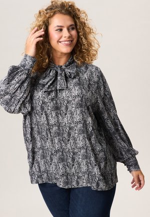 Lachende vrouw met krullend haar, gekleed in een zwart-witte blouse met slangenprint en strikceintuur, en een donkere spijkerbroek, poserend tegen een effen achtergrond.