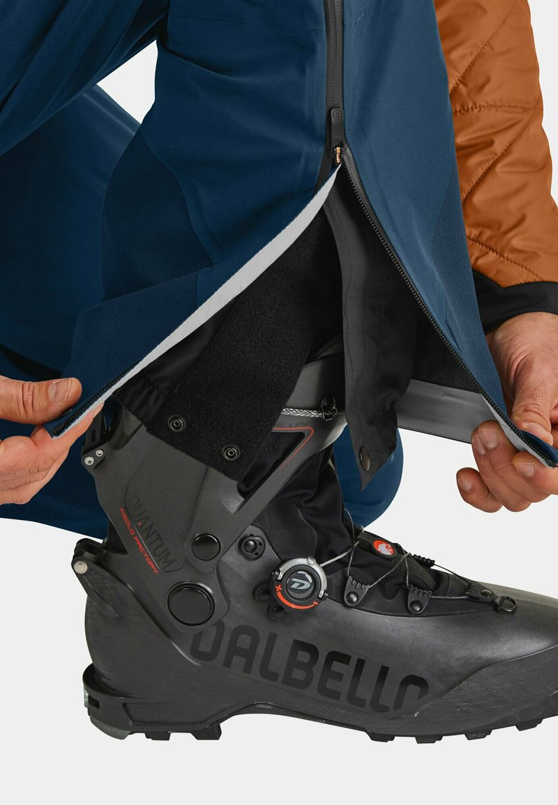 Schwarzer Skischuh mit Dalfello-Branding, platziert neben dunkelblauen Skihosen. Die Hosen verfügen über einen Reißverschluss am Saum für einfachen Zugang.