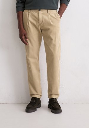 Beige corduroy broek met plooien, rechte pijpen en textuurstof. Gecombineerd met donkerbruine suède schoenen voor contrast.