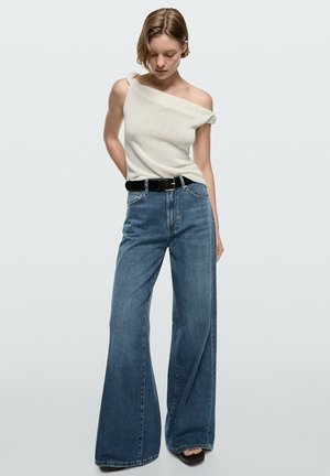 Femme portant un top blanc asymétrique, un jean bleu à jambes larges avec une ceinture noire et des chaussures noires à bout ouvert, debout devant un fond uni.