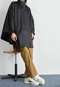 Svart oversize poncho med hög krage, gjord av ett mjukt material. Bärs över beige byxor och vita sneakers. Enkel design utan synliga mönster.
