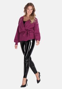 Lila sálkötött kardigán gyöngyökkel díszített vállakkal, derékban megkötve; fényes fekete leggings-szel és fekete magassarkú cipőkkel párosítva.