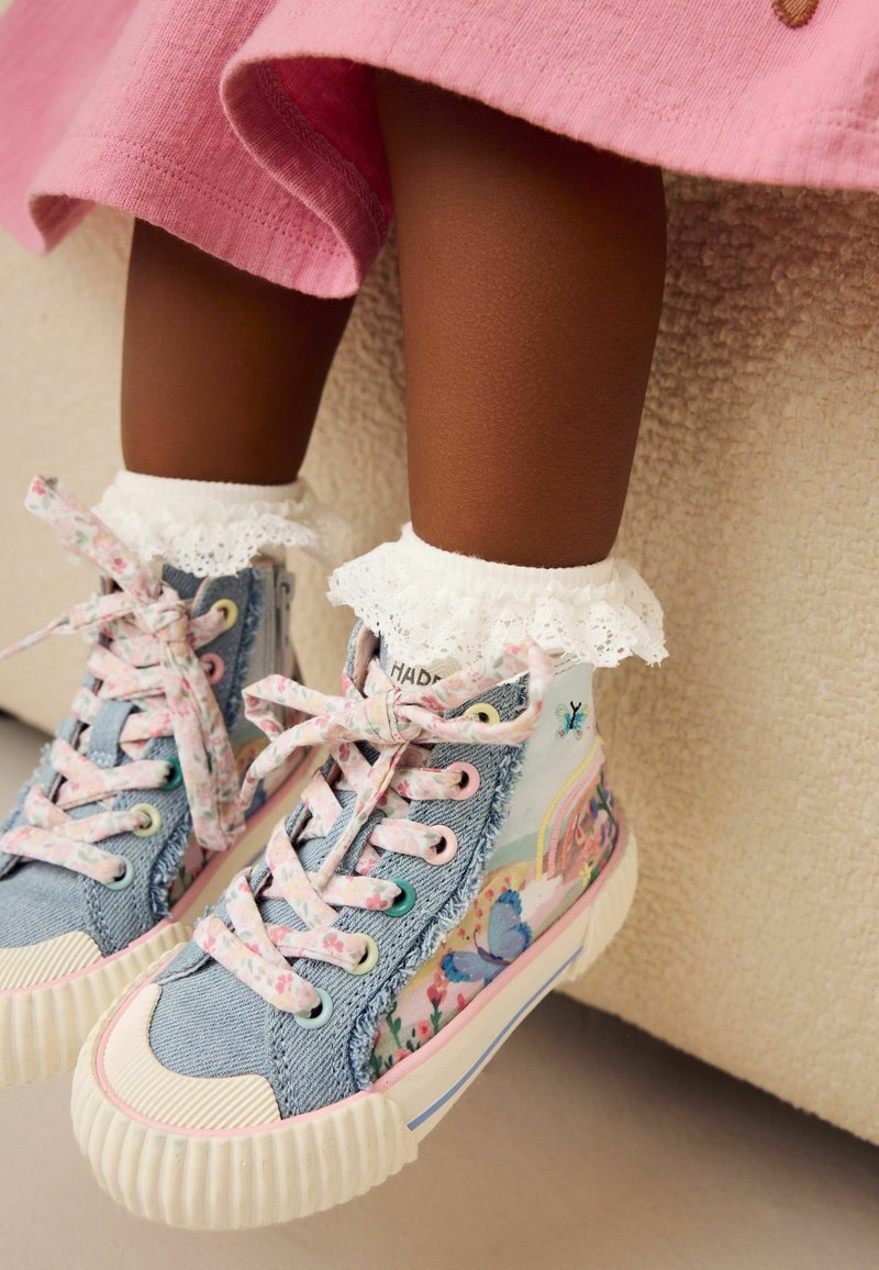 Jambes d'enfant portant une robe rose, des chaussettes blanches à volants et des baskets montantes bleues avec des motifs floraux et papillons, pendant au-dessus d'une surface beige.