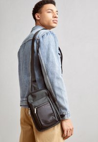 Borsa a tracolla nera e grigia con tasche con zip, forma rettangolare e tracolla regolabile, indossata sopra una giacca di jeans e pantaloni beige.