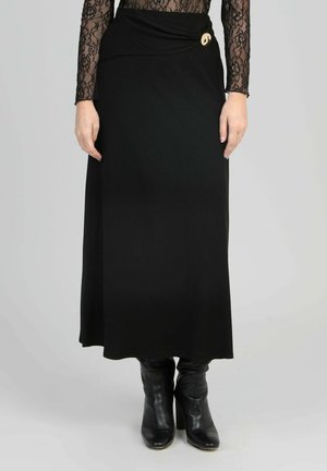 Femme portant une longue jupe noire avec un bouton décoratif doré et des bottes en cuir noires à talons, associées à un haut à manches longues en dentelle noire.