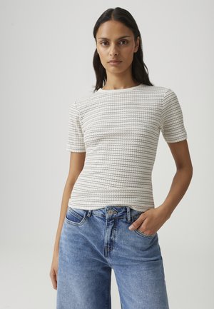 Vrouw met lang donker haar die een wit gestructureerd kortermig topje en blauwe hooggetailleerde spijkerbroek draagt, staand tegen een effen achtergrond.