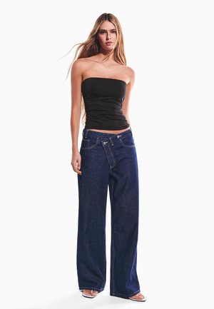 Ung kvinde med langt hår iført en sort stropløs top, oversized mørkeblå jeans og hvide åbne hæle, stående mod en hvid baggrund.