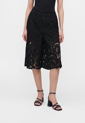 Γυναίκα με μαύρα φλοράλ δαντελωτά culottes και μαύρα σανδάλια με λουράκια και χοντρό τακούνι, στέκεται μπροστά σε μια απλή φόντο.