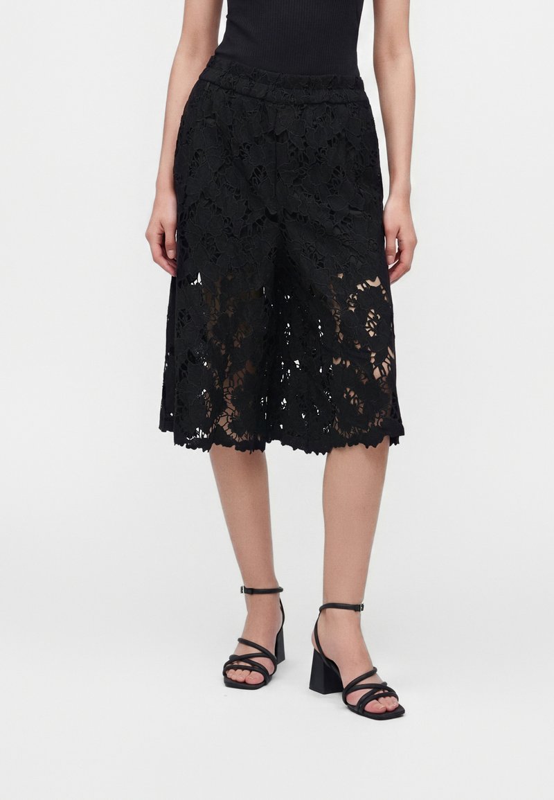 Γυναίκα με μαύρα φλοράλ δαντελωτά culottes και μαύρα σανδάλια με λουράκια και χοντρό τακούνι, στέκεται μπροστά σε μια απλή φόντο.