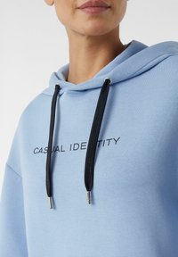 Felpa azzurro chiaro con cappuccio, dotata di un cordino nero, componenti in metallo argentato e testo stampato "CASUAL IDENTITY" sul davanti. Tessuto liscio e strutturato.