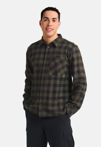 Chemise en flanelle à carreaux vert foncé et bleu marine, avec une fermeture à boutons, des manches longues et une poche sur la poitrine. Le tissu a une texture douce.