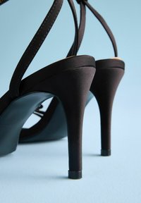 Schwarze Stiletto-Absätze mit Satinriemen, die eine glatte Textur und einen zugespitzten Absatz bieten. Die Sohle ist matt schwarz mit einem leichten Glanz.