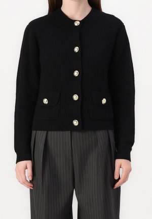 Person wearing a black ribbed cardigan with six gold buttons and two flap pockets, paired with dark gray pinstripe trousers.