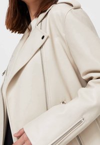 Cremefarbene Kunstlederjacke mit einem diagonalen Reißverschluss, breitem Kragen und Reißverschlussmanschetten. Glatte Textur mit minimalistischen Designelementen.