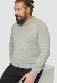 Grauer Pullover mit strukturiertem Strick, Rundhalsausschnitt und gerippten Bündchen, mit dunklenFlecken für zusätzliche Details. Getragen über schwarzen Hosen.