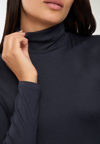 Kobieta poprawiająca kołnierzyk granatowego golfu, odsłaniająca usta, brodę i część swoich długich ciemnych włosów.