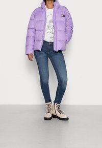 En person iklädd en lavendel puffjacka, vit t-shirt med grafik, blå skinny jeans och beige snörboots står på ett grått golv mot en vit vägg.