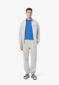 Chaqueta gris con cremallera, camiseta azul, pantalones deportivos grises y sandalias deslizables azules. La tela parece suave; incluye detalles del logo en las prendas.