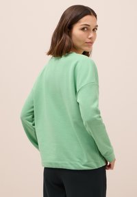 Sudadera verde claro con un corte relajado y mangas largas, que presenta un escote redondo y un dobladillo recto. La tela parece suave con una textura mínima.