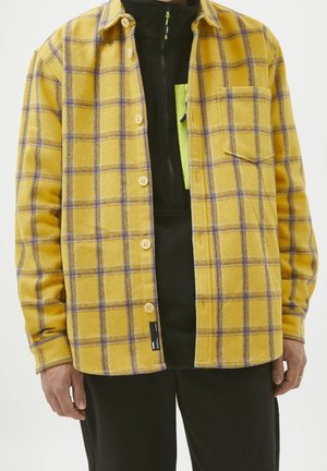 Personne portant une chemise à carreaux jaune et violette boutonnée par-dessus un haut noir zippé avec un détail de poche poitrine vert fluo.